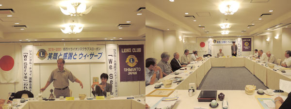 ホテルの会議室で音次郎の話をする浦田会長と事務局の瀬戸。新しい伝記を掲げている。ロの字型に会議用テーブルが並び、それを取り囲んで12人のライオンズメンバーが話を聞いている。