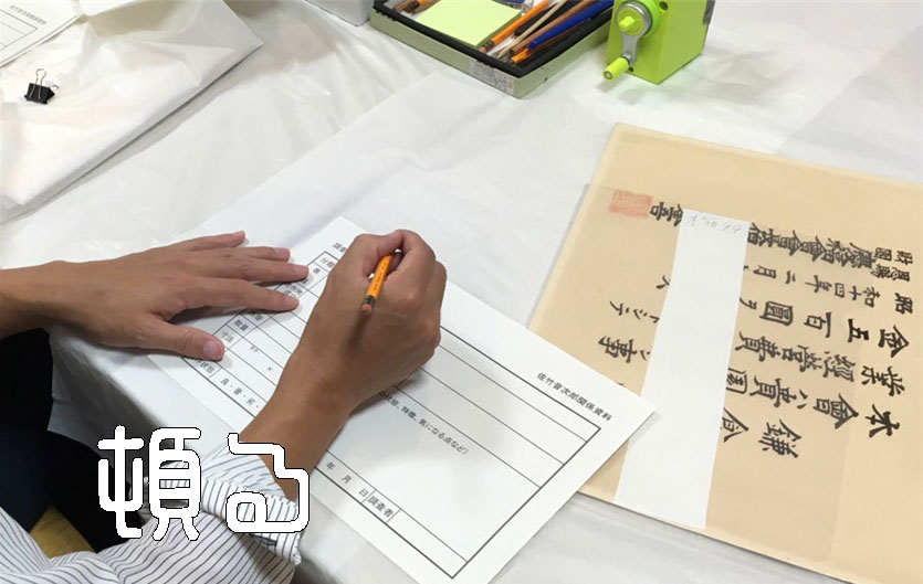 何かの表彰状を右に置いて鉛筆を握り調査票に書き込んでいる左右の手の写真。史料整理作業の１コマ