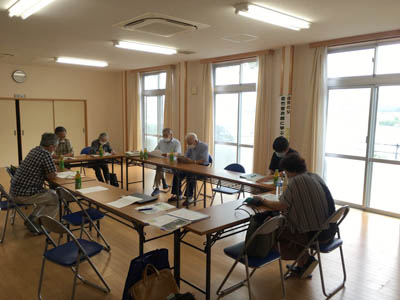 竹島防災センターの室内に会議用テーブルをロの字に並べて中平会長の話に聞き入る人たち。写真の後ろの窓は明るいが大雨が降っている。