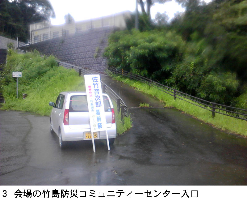 会場の竹島防災コミュニティーセンター入口。軽自動車のトランクハッチに墓前祭会場を記す看板が固定されている。きつい坂道を上がった上に会場はある