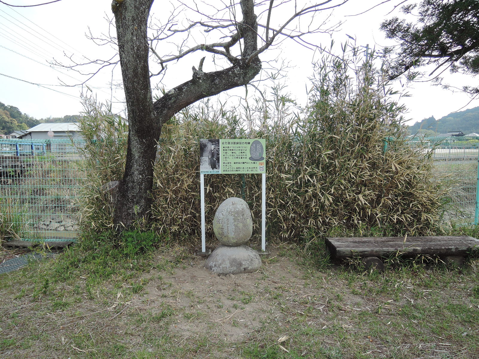 竹島集会所の川沿いフェンス前、左に栴檀の木、背後に笹林、右には丸太風ベンチ。緑字に説明文と鎌倉の歌碑の写真が入った案内板。