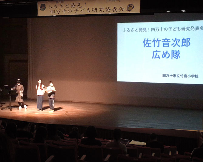 舞台の上に立つ竹島小５年生３人。上には「第３回 ふるさと発見！四万十の子ども研究発表会」の看板がある。１人の男児が右後ろを振り返り、スクリーンに映された「佐竹音次郎　広め隊」の文字を見ている。