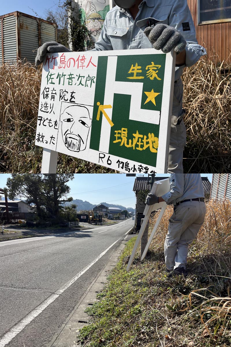 竹島小学校の令和６年度卒業生が工夫して描き、完成した木製看板。今まさに看板屋の手によって据え付け工事がなされようとしている所。作業服姿に軍手、斜面に看板をしっかり支えながら立つアオキ工芸さん。