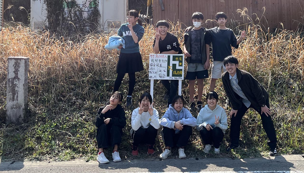 完成した看板と並んで記念撮影。担任の教師と、児童８人が看板を取り囲む。男子３名、女子５名。ピースをしている子供や、斜めを向いてポーズを取るおませさんも居る。